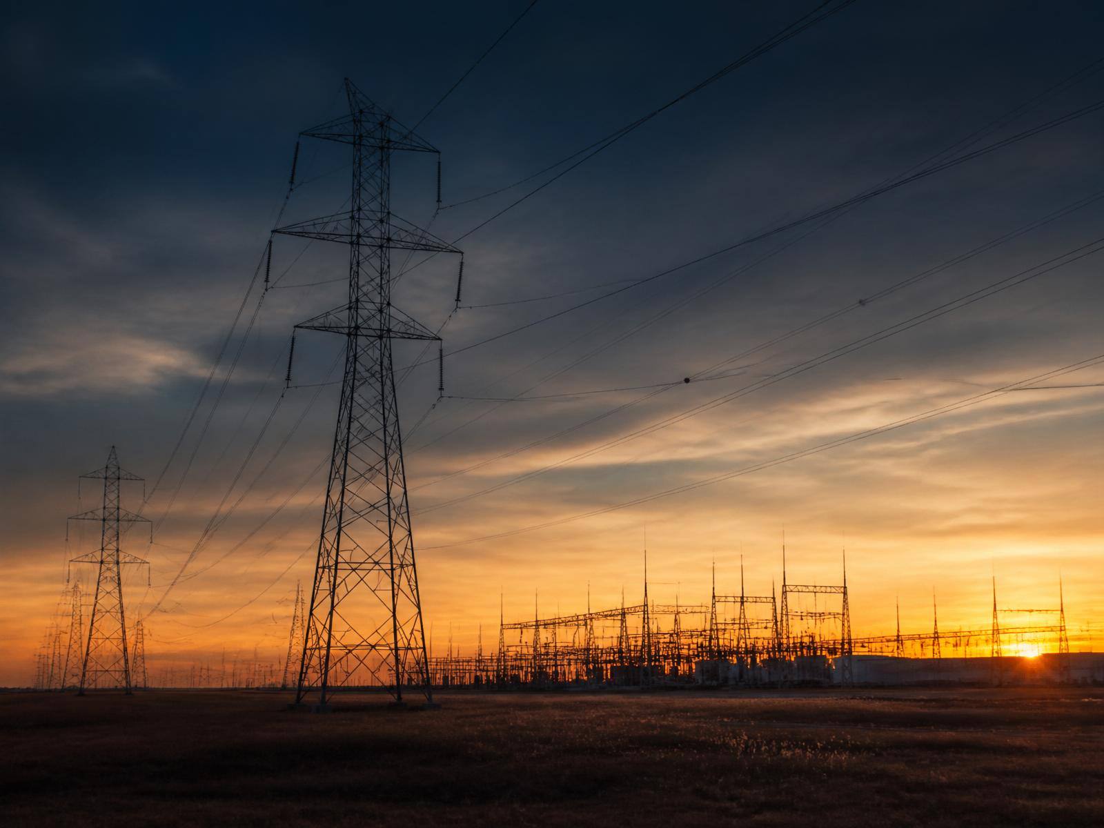 High-voltage transmission pylons and a substation silhouetted against a warm sunset sky.